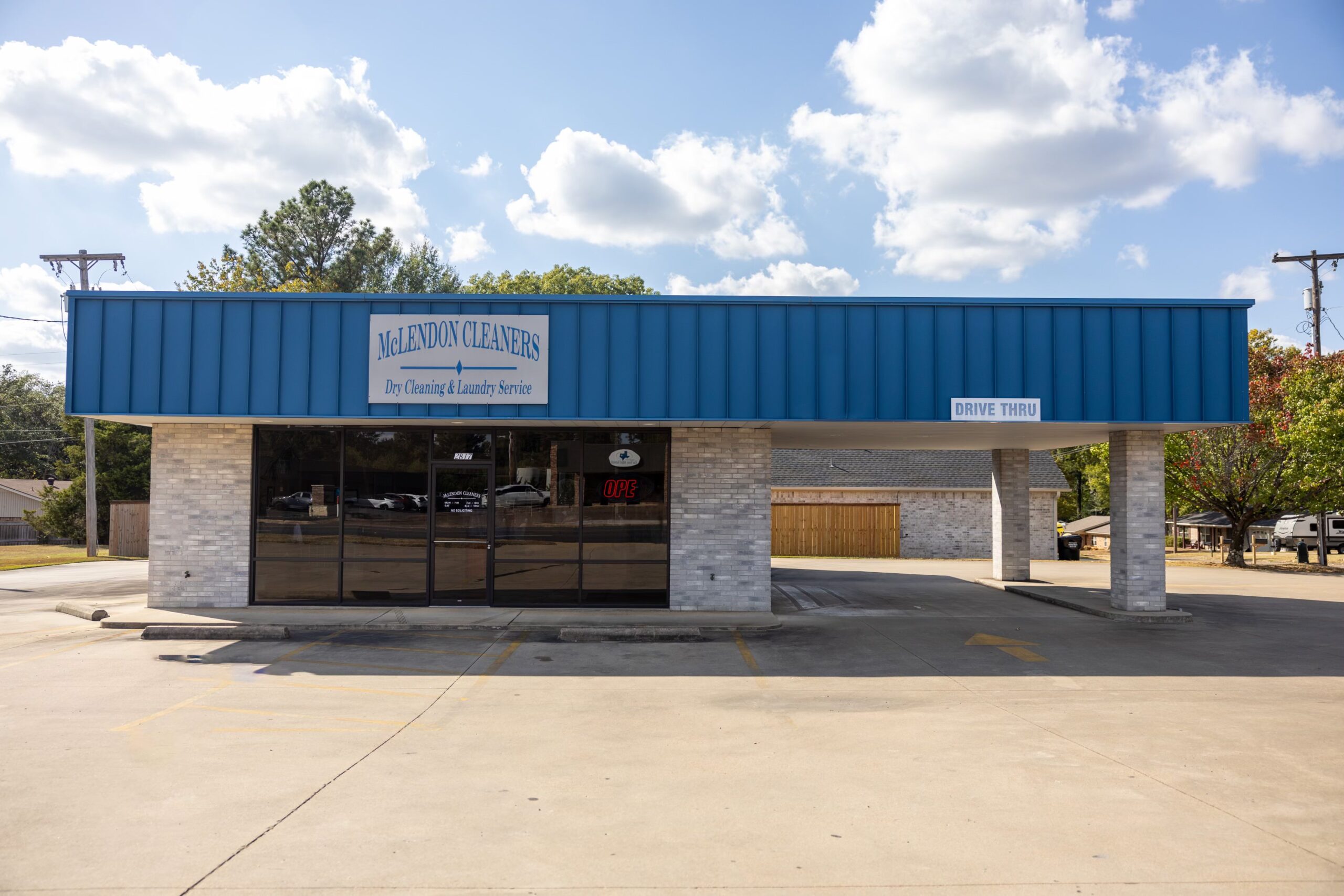 A dry cleaning and laundry service building with a drive-thru, featuring a blue sign and brick exterior under a blue sky with clouds.