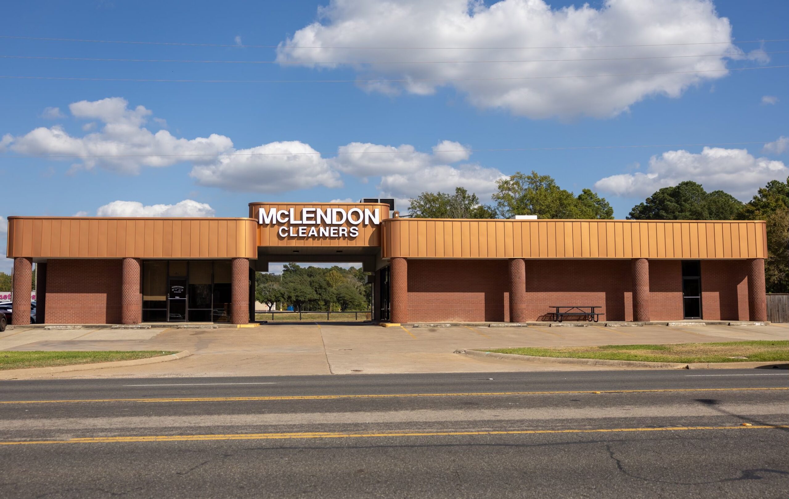 A brick building with the sign "McLendon Cleaners" is located by the roadside, under a partly cloudy sky.