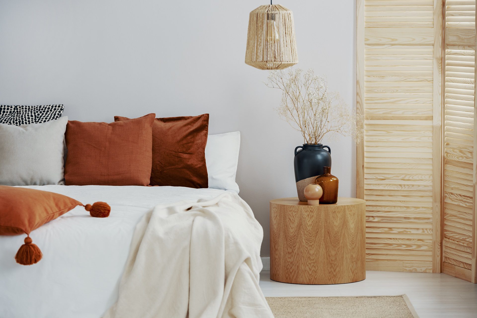 Bedroom with a neatly made bed featuring brown and white pillows, a beige blanket, a wooden nightstand with decorative vases, and a rattan pendant lamp.