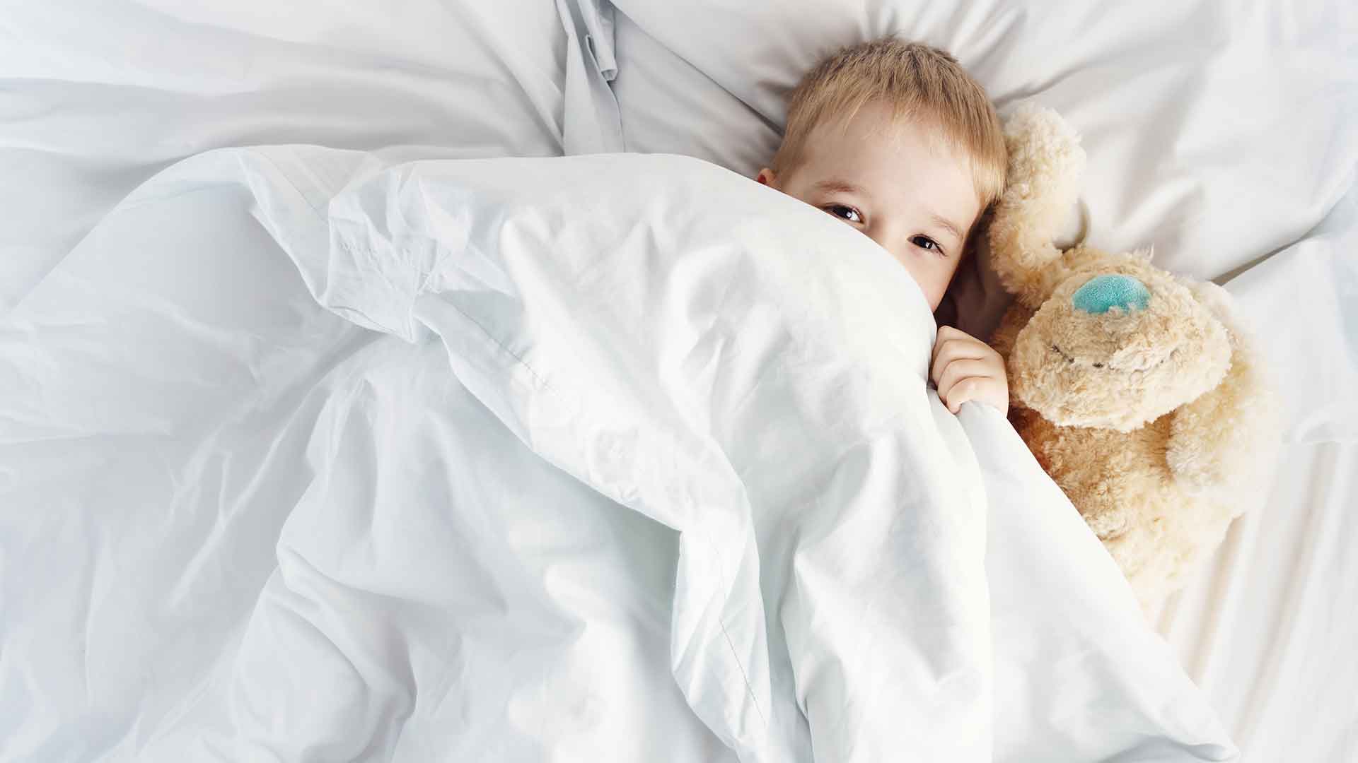 A child lies in bed under a white blanket, holding a teddy bear.