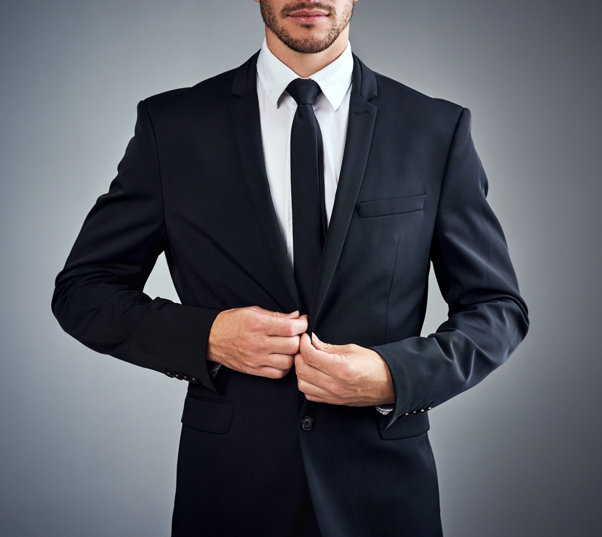 A person in a black suit adjusts their jacket against a neutral background. Their face is not visible.