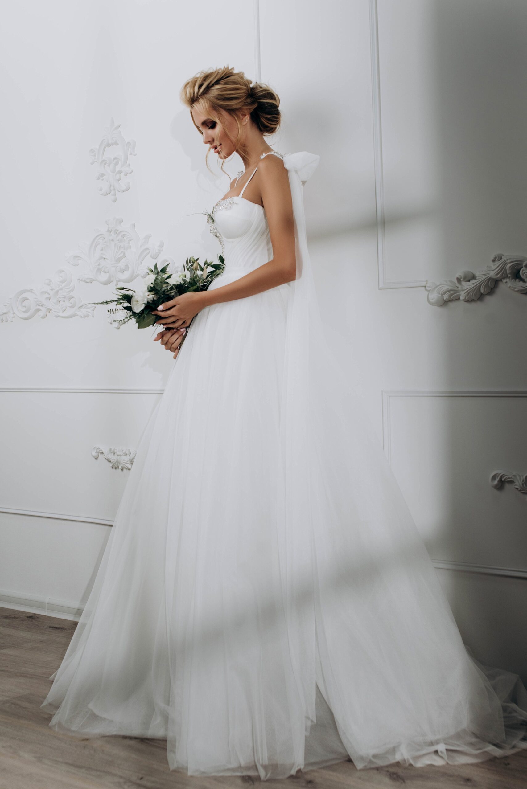 A woman in a white wedding dress holds a bouquet, standing in a softly lit room with elegant white wall detailing.