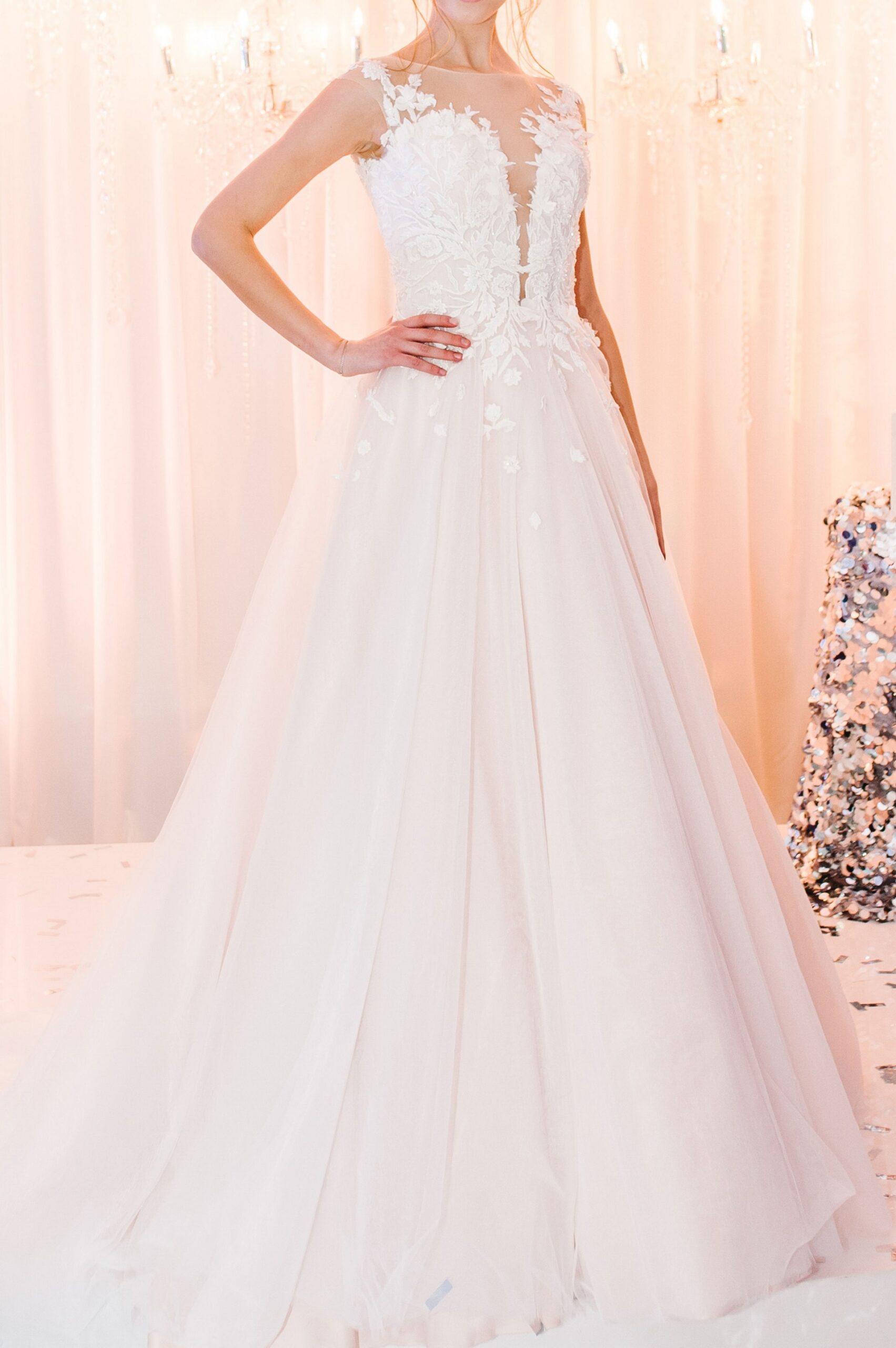 A bride wearing a white lace wedding gown with floral details and a tulle skirt stands with one hand on her hip in front of a softly lit background.