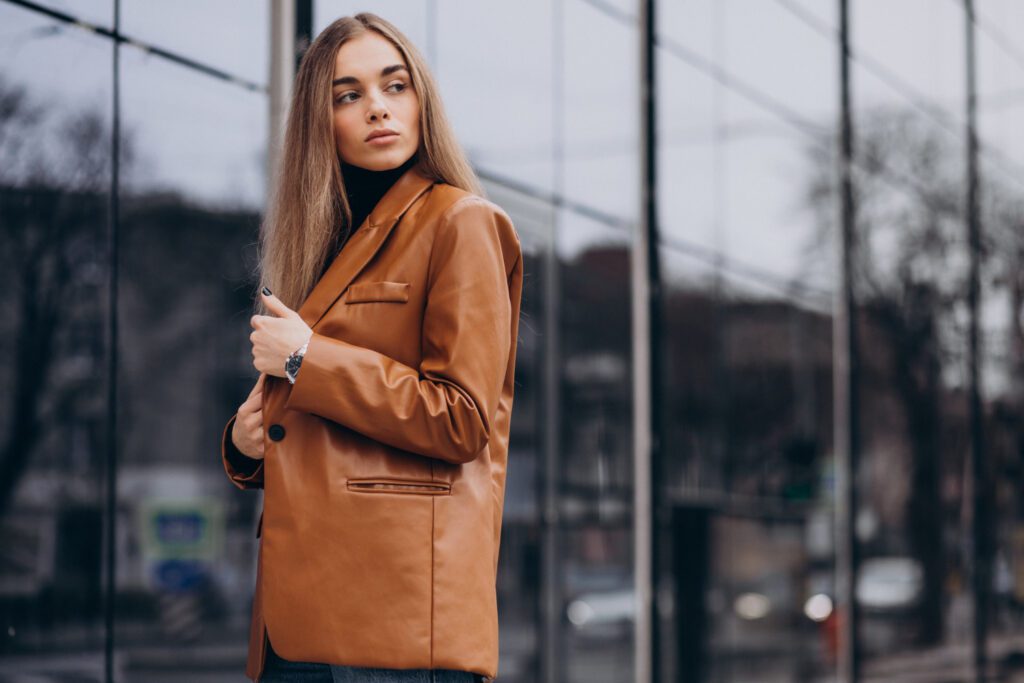Woman with long hair wearing a brown leather jacket stands in front of a reflective glass building, looking to the side.