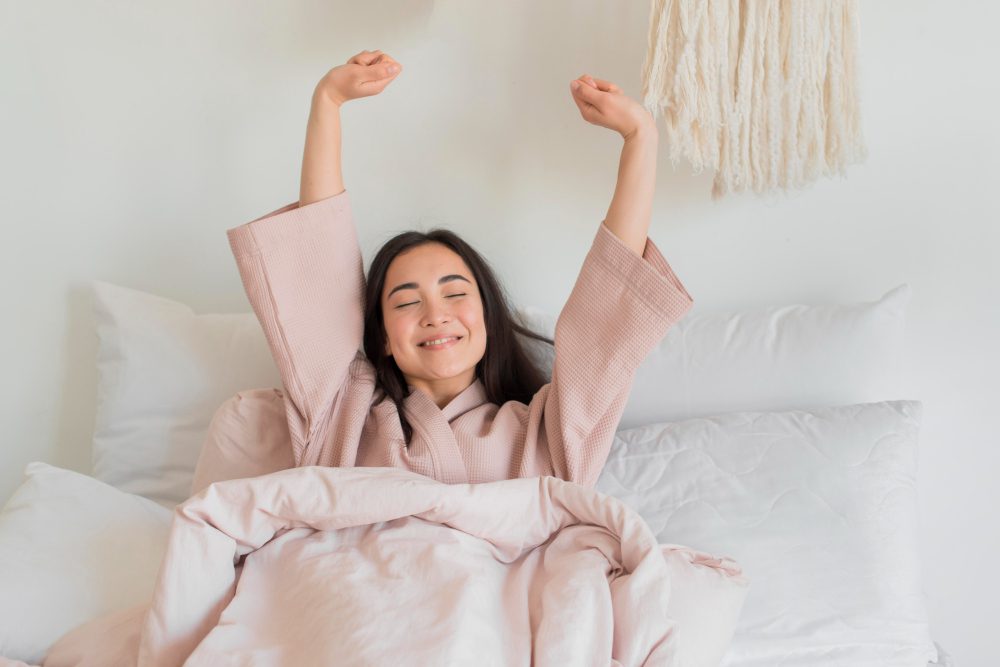 A woman in a pink robe sits up in bed, smiling with eyes closed and arms stretched overhead, surrounded by white pillows and bedding.