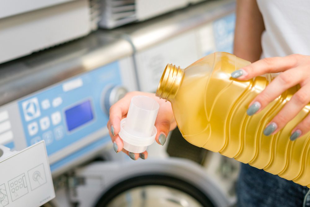 A person pours liquid laundry detergent from a yellow bottle into a measuring cap in front of a washing machine.