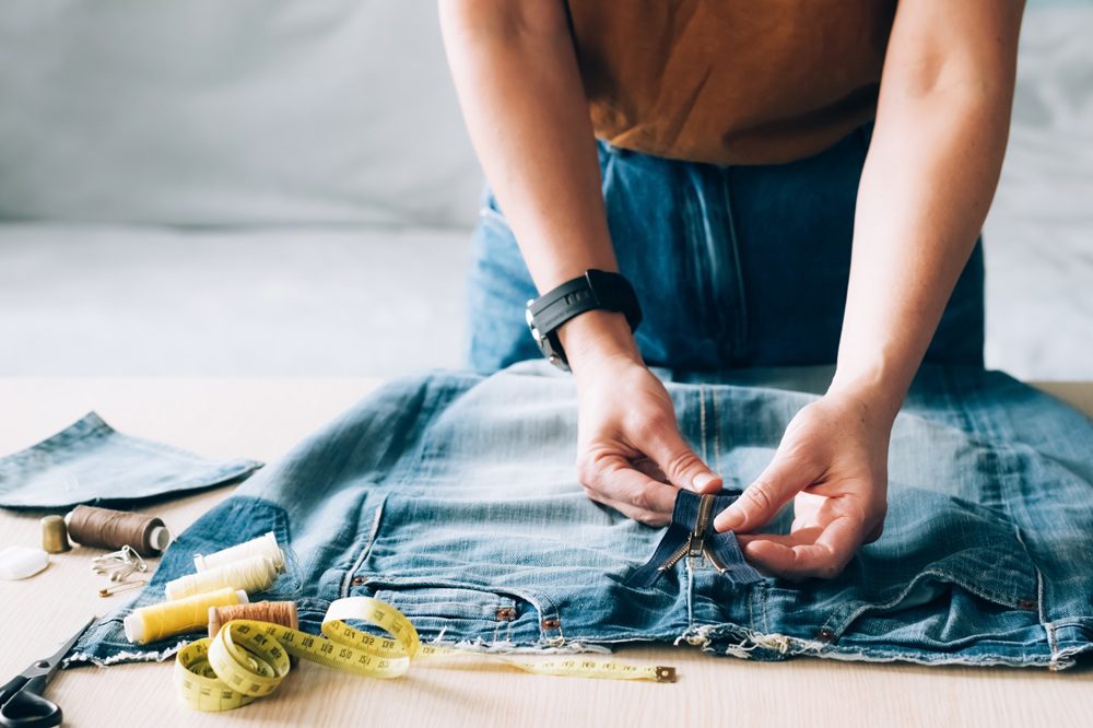 A person works on a pair of jeans at a table with sewing materials, thread spools, scissors, and a measuring tape.