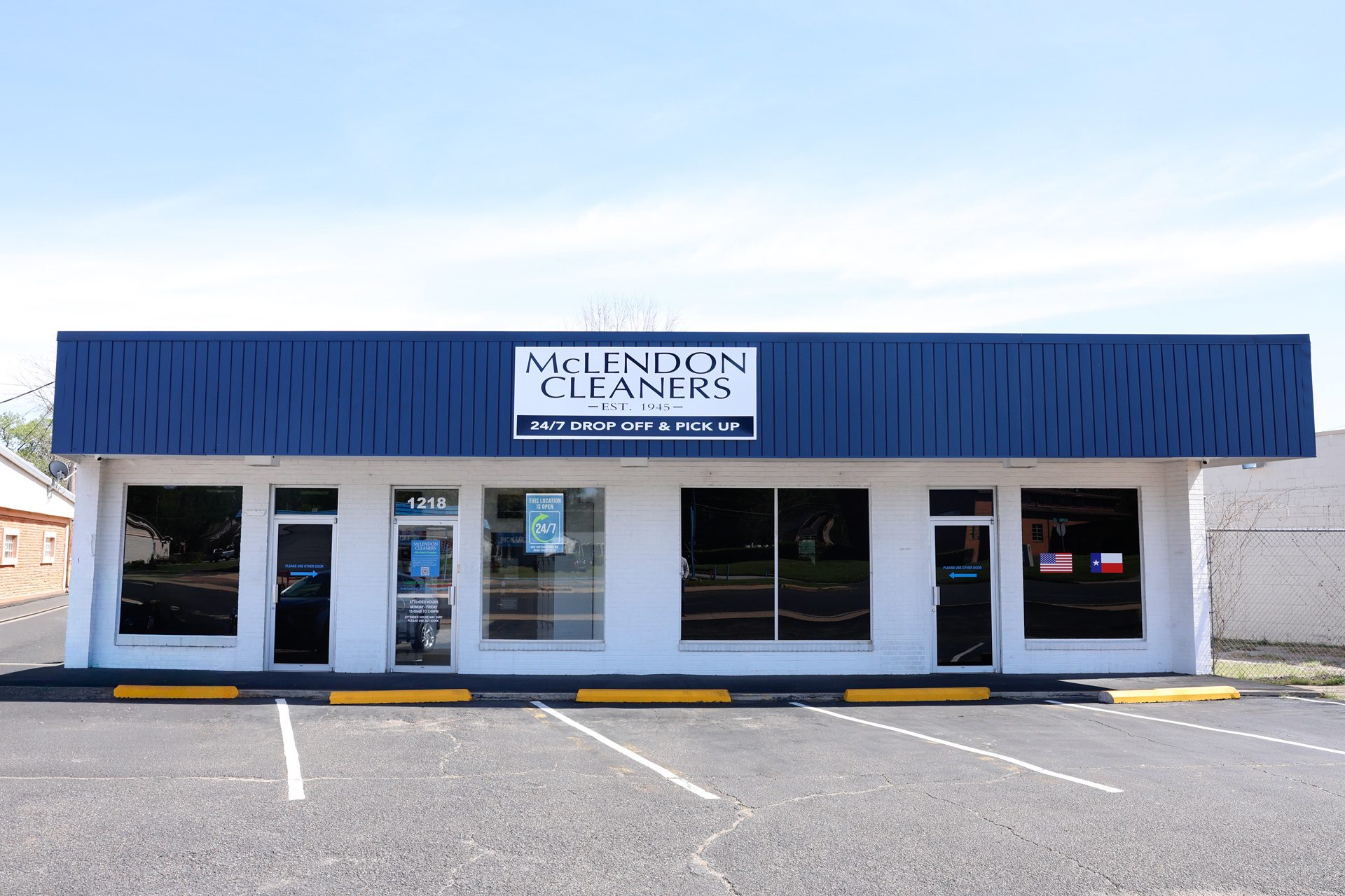 A white and blue building with a sign reading "McLendon Cleaners," featuring large windows, a glass door, and an empty parking lot in front.