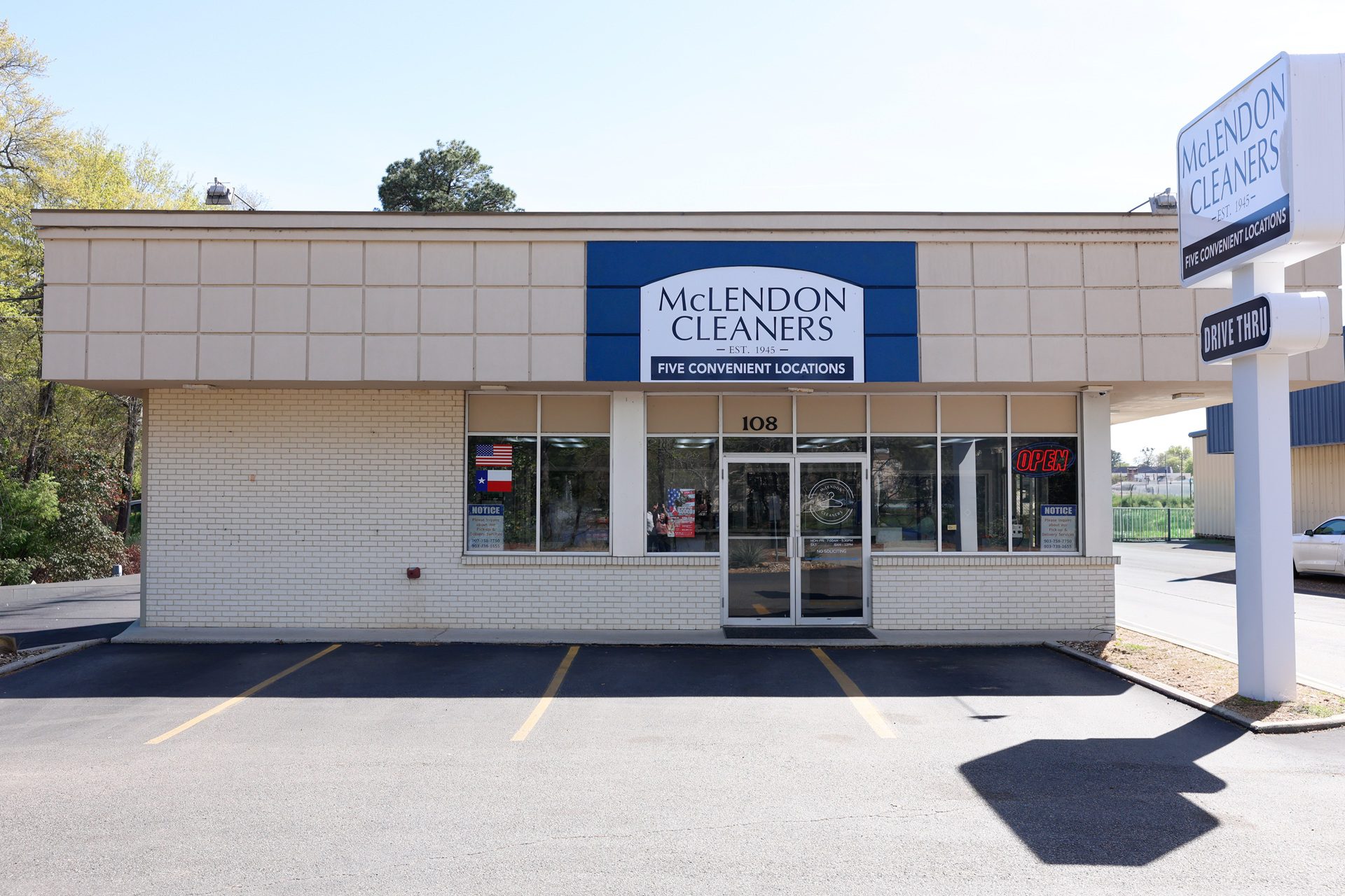A single-story building with a "McLendon Cleaners" sign above the entrance, clear windows, an "OPEN" sign, and an empty parking lot in front.