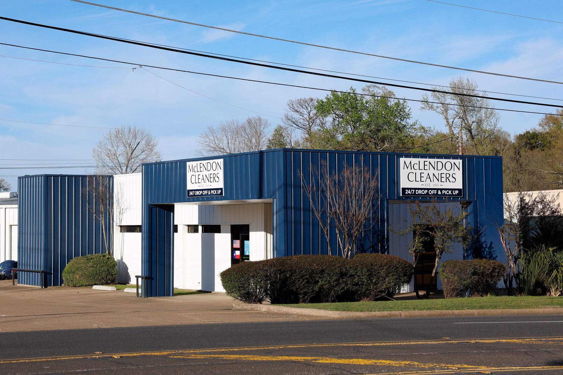 A blue and white building with signs reading "McLendon Cleaners" and "24/7 Drop Off & Pick Up," surrounded by bushes along a street.